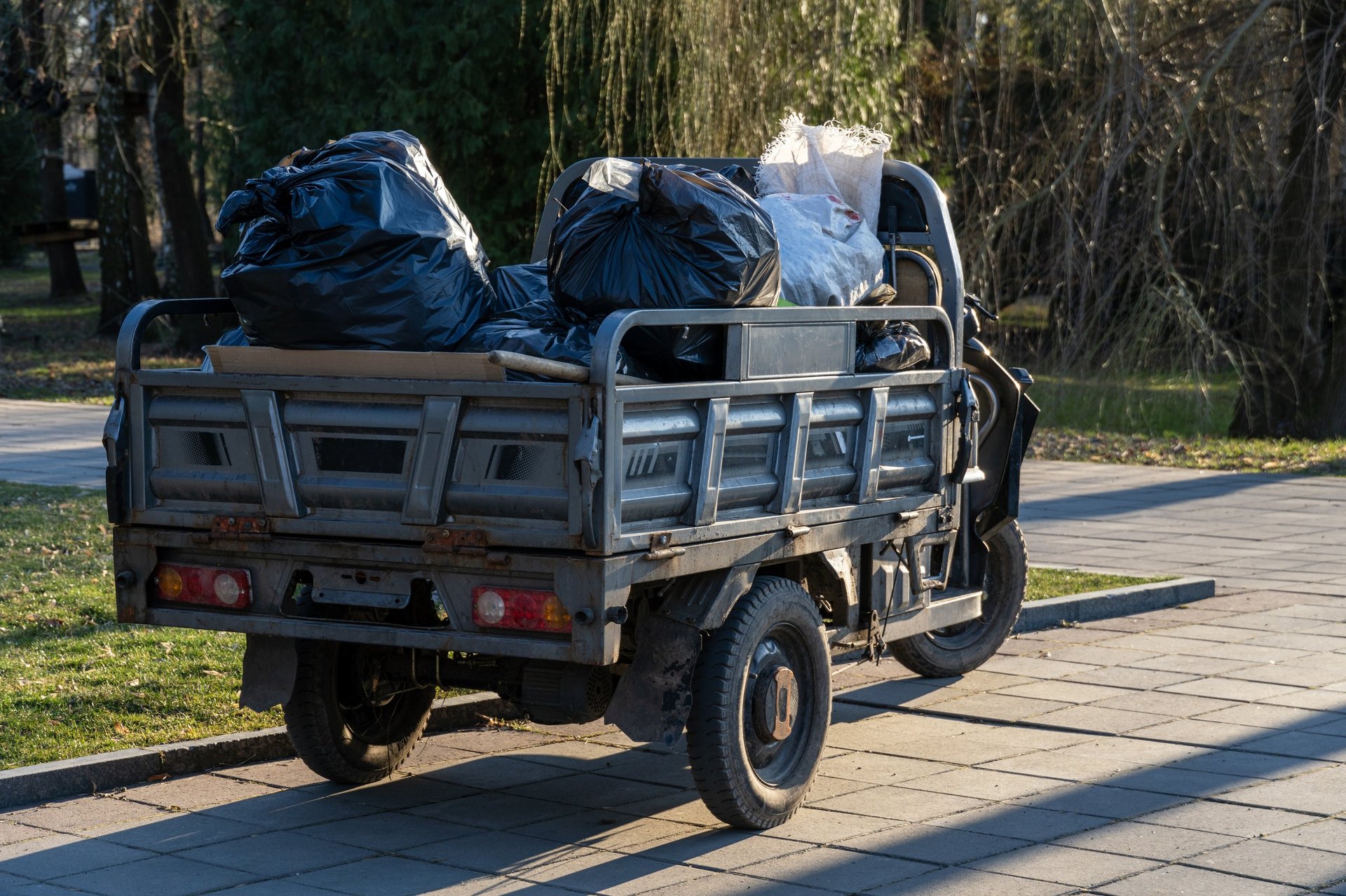 A small truck filled with black garbage bags parked along a city pathway, showing junk removal pickup