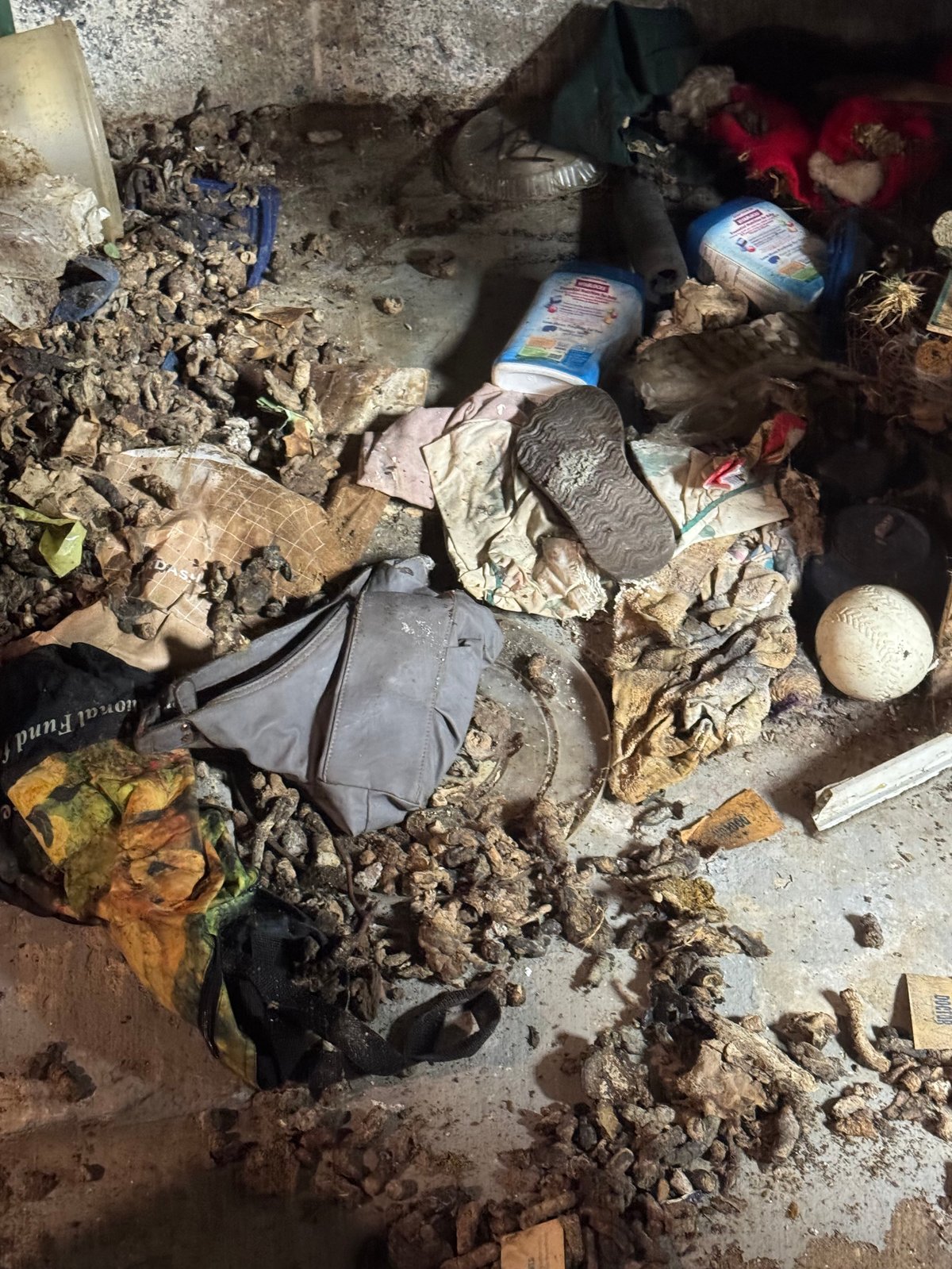 Overhead view of cluttered ground with scattered trash, plastic bags, concrete debris, and personal items among dirt and rubble