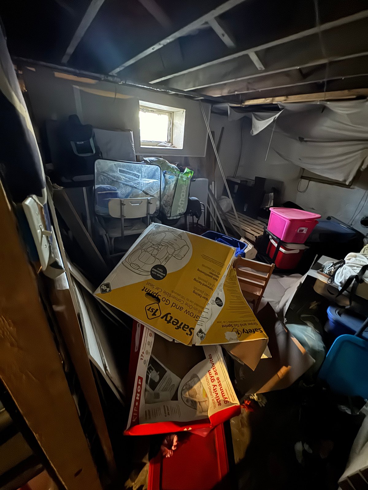 Cluttered garage or storage space with cardboard boxes, plastic containers, and miscellaneous items scattered on the floor and shelves.